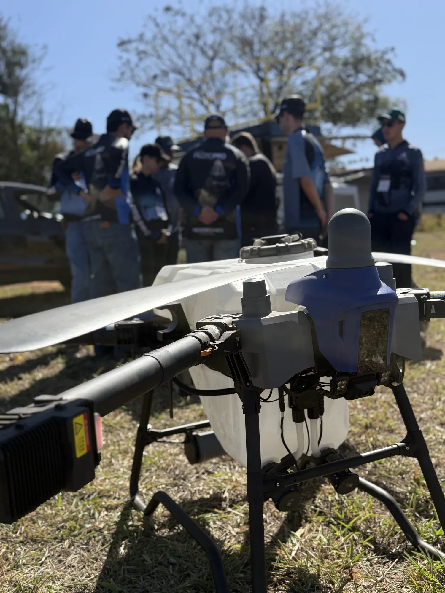 Drone agrícola XAG no campo de voo, com a turma da Imersão Piloto PRO ao fundo